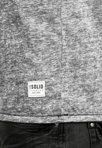 Graues Melange-T-Shirt mit abgerundetem Saum, versehen mit einem Etikett mit "SOLID" und dem Gründungsdatum. Weicher Stoff mit sichtbaren Nähdetails.
