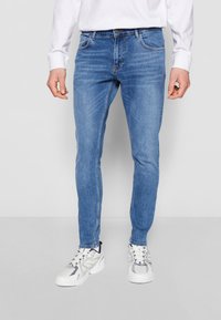 Vaqueros de denim azul de corte ajustado con un lavado intermedio, diseño de cinco bolsillos, cierre de botón y sutiles detalles de costura, combinados con zapatillas blancas.