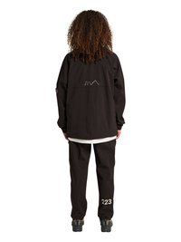 Person med krøllet hår iført en sort jakke med en bysilhuet på ryggen og sorte bukser mærket "223" på venstre ben.