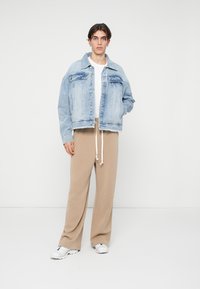 Vaate denim-takki vaaleansinisessä värissä, jossa on rento malli, nappikiinnitys, taskupusseja ja kulunut helma, yhdistettynä beigeihin leveälahkeisiin housuihin.