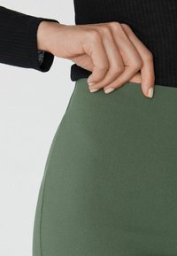 Kalhoty s vysokým pasem, olivové zelené barvy, vyrobené z texturované látky, se švy podél stran a přiléhavým střihem.