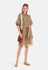 Kleid aus khakifarbenem Leinen mit Puffärmeln, einem Ausschnitt am Hals und einer gebundenen Taille. Kombiniert mit einer braunen Umhängetasche und gewebten beigen Ballerinas.