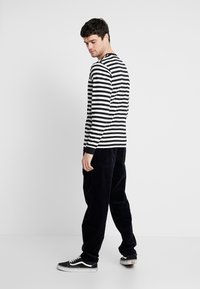 Cămașă cu dungi verticale alb-negru, cu mânecă lungă, asortată cu pantaloni negri și adidași negri, având o croială lejeră și elemente de design simple.