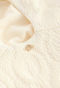 Texturiertes weißes Gewebe mit erhabenen Mustern und einem kleinen, goldfarbenen Verschluss. Das weiche, glatte Innenmaterial ist unter dem Gewebe sichtbar.