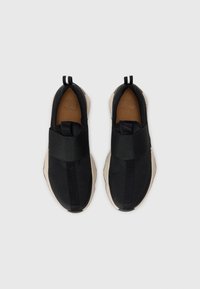 Par svart slip-on sneakers med elastisk stropp og beige såler sett ovenfra på hvit bakgrunn.