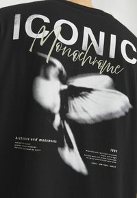 Czarna koszulka z dużym monochromatycznym wzorem kwiatu, z tekstem "IKONICZNY" i "Monochrom" powyżej oraz dodatkowymi detalami poniżej.