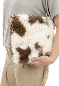 Borsa a tracolla in pelliccia sintetica con stampa a pois di mucca marrone e bianca. Presenta una tracolla color sabbia e un logo impresso su una linguetta in pelle. Texture morbida, forma rettangolare.
