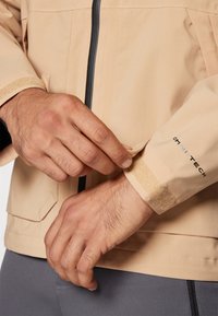Beige wasserdichte Jacke mit schwarzem Reißverschluss und Velcro-Manschetteneinstellung. Verfügt über eine glatte Textur und ergonomischen Schnitt. Logo am Ärmel.
