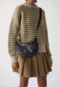 Persona che indossa un maglione a righe in maglia e una gonna plissettata color beige, mentre porta una borsa a tracolla a forma di mezzaluna in pelle nera con una tracolla in catena argentata.