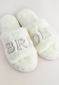White fur-lined slippers featuring "BRIDE" adorned with pearl accents and rhinestones on a soft, textured strap. Rounded shape and cushioned sole.