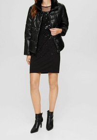 Černá péřová bunda s límcem z umělé kožešiny, přes černé šaty s flitry. Modelka má na sobě černé kozačky nad kotník s špičatou špičkou.