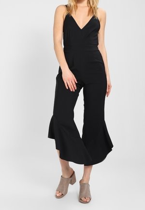 Czarny kombinezon z dekoltem w kształcie litery V, dopasowanym gorsetem i rozszerzanymi, skróconymi nogawkami. Tkanina o strukturze; zestawiony ze szarymi, jasnymi, odkrytymi butami na obcasie.