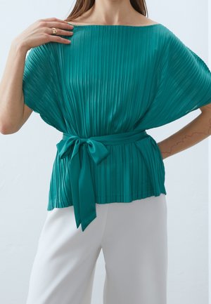 Blusa plissettata teal con maniche larghe, scollo off-the-shoulder e dettaglio della vita legato. Abbinata a pantaloni bianchi su misura.