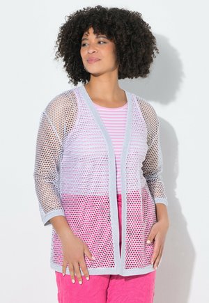 Vrouw met krullend haar die een witte opengewerkte cardigan draagt over een roze gestreepte top en felroze broek, staand tegen een witte muur.