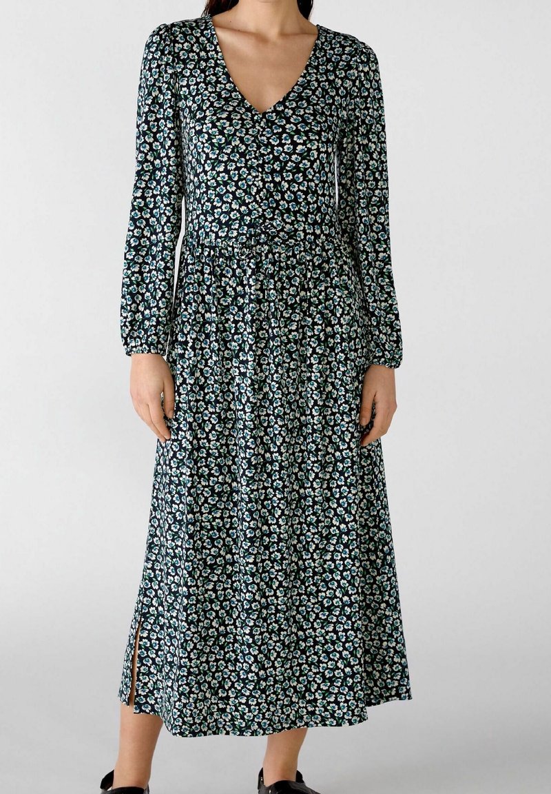 Femme portant une robe midi à manches longues avec un motif floral bleu et blanc, décolleté en V, taille froncée et fente latérale sur fond bleu marine.