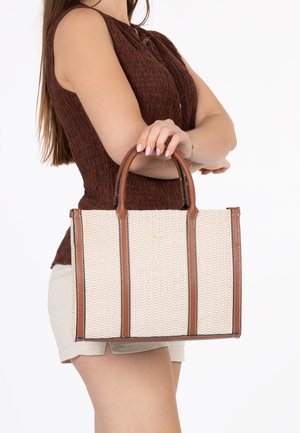 Femme portant un haut marron sans manches et un short beige tient un sac fourre-tout tissé beige avec des poignées et des bordures en cuir marron.