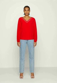 Sweater vermelho de malha com decote em V e rendas, combinado com jeans retos de perna clara e sapatos de salto aberto brancos.