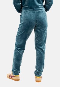 Personne portant un pantalon en velours teal et des baskets beiges avec des rayures rouges, se tenant avec les mains dans les poches, vu de dos.