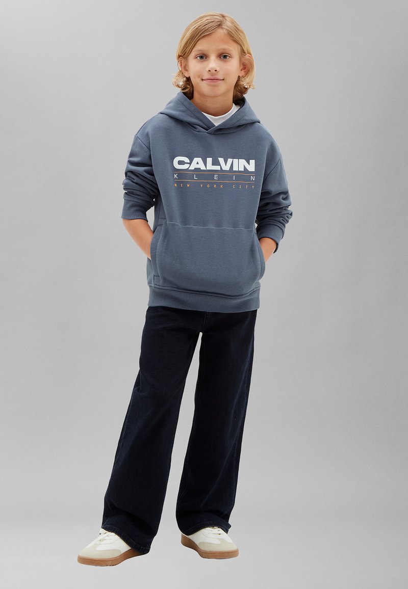 Enfant aux cheveux blonds portant un sweat à capuche bleu Calvin Klein, un pantalon noir et des baskets blanches, debout les mains dans les poches sur fond gris.
