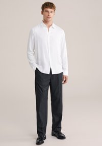 Camicia bianca con bottoni e maniche lunghe, abbinata a pantaloni grigi scuri su misura e scarpe nere, in piedi su uno sfondo neutro.