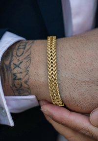 Goldene Kettenarmband mit einem geflochtenen Design, das das Licht reflektiert. Getragen an einem Handgelenk mit kurzem, feinem Haar und einem sichtbaren Tattoo darunter.