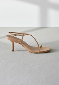 Beige hoge hakken sandalen met een dunne bandontwerp, vierkante tenen en een lichte glanzende afwerking. Kenmerkt een delicate enkelband met een gesp.