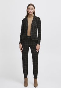 Un ensemble de costume à carreaux noir et doré avec une veste ajustée et un pantalon fuselé, associé à un pull col roulé beige et des bottines beiges.