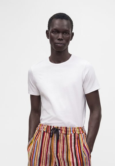 Personne à la peau foncée portant une chemise blanche unie à manches courtes et un pantalon rayé coloré, les mains dans les poches, sur fond blanc.