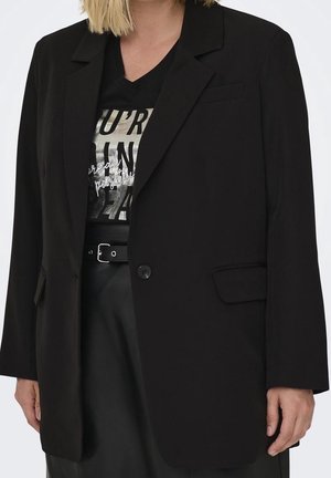 Kobieta ubrana w czarną marynarkę na czarnym T-shircie z grafiką oraz czarne spodnie z paskiem, widoczna od ramion do bioder.