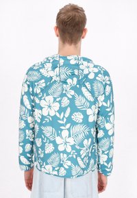 Huvtröja med en turkos bakgrund och stora vita blommor och bladmönster. Tillverkad av lätt, slät tyg.