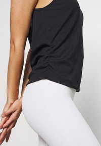 Débardeur noir côtelé avec un détail froncé sur le côté, associé à des leggings blancs ajustés, présentant une texture lisse et une coupe près du corps.