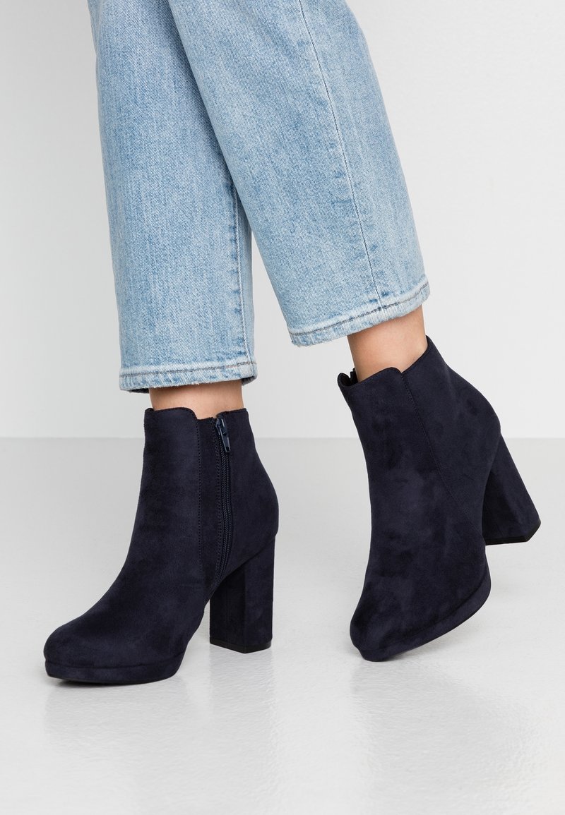 Personne portant un jean court bleu clair et des bottines en daim noir à talons blocs avec des fermetures éclair latérales sur un sol blanc.