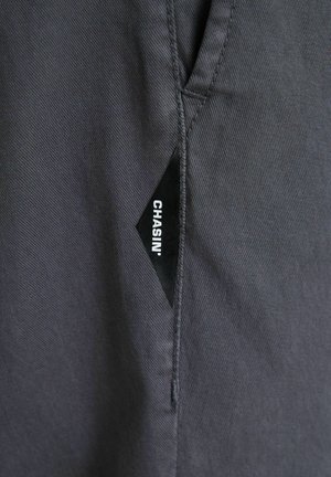 Etichetă de țesătură în formă de diamant de culoare neagră cu text alb „CHASIN'” cusută în cusătura unei piese de îmbrăcăminte gri închis.