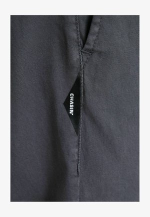 Etichetă de țesătură în formă de diamant de culoare neagră cu text alb „CHASIN'” cusută în cusătura unei piese de îmbrăcăminte gri închis.