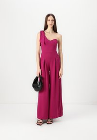 Fuksia ensidig jumpsuit med plissert vide ben, glatt tekstur, og en sløyfeaccent. Kombinert med en svart håndveske og sandaler med stropper.