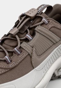 Καφέ αθλητικό παπούτσι με υφή επάνω μέρος, ελαστικά κορδόνια, καουτσούκ σόλα και ασημί λεπτομέρειες. Διαθέτει επενδεδυμένο κολάρο και έντονο λογότυπο στο πλάι.