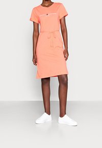 Koralli lyhythihainen mekko, jossa on pyöreä pääntie, säädettävä nyörivyötärö ja logoteksti. Yhdistetty valkoisiin tennareihin neutraalilla taustalla.