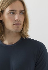 Maglietta a maniche lunghe blu navy con scollo tondo, texture liscia e sottile lucentezza. Inquadratura ravvicinata della parte superiore del torso e del volto.