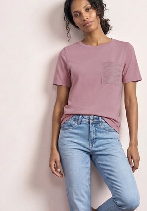 Kobieta ubrana w wrzosową koszulkę z krótkim rękawem z wzorzystą kieszenią i jasnoniebieskie dżinsy, stojąca na tle jednolitej jasnej ściany.