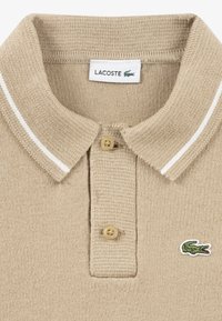Beige polo shirt with a textured knit, two-button placket, and contrasting white stripes on the collar. Features a green Lacoste logo.