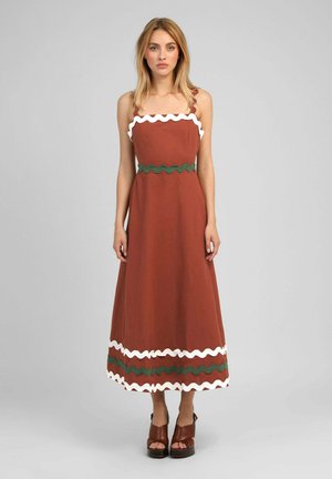 Femme debout portant une robe midi sans manches couleur rouille avec une bordure ondulée blanche et verte et des sandales à plateforme marron.