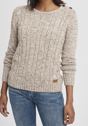 Kvinde iført en beige kabelstrikket sweater med knappedetaljer på skulderen og blå jeans, stående mod en ensfarvet baggrund.