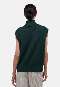 Pull sans manches vert foncé à côtes avec un col haut, en tissu tricoté texturé, et une silhouette décontractée, associé à un pantalon rayé clair.