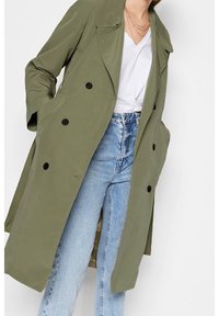 Trench de color verde oliva con un diseño de doble botonadura, botones negros, solapas anchas y dos bolsillos laterales sobre unos jeans de color azul claro.