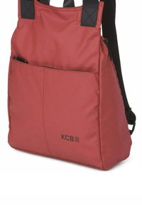 Mochila de tela roja con correas negras, que cuenta con un bolsillo frontal con cremallera y un logo en la esquina inferior derecha. Textura suave y diseño minimalista.