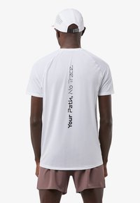 Weißes Sportshirt mit strukturiertem Oberflächenmaterial, auf dessen Rückseite ein vertikales Design aus schwarzem Text zu sehen ist. Kombiniert mit hellbraunen Shorts.