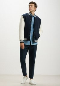 Giacca varsity blu navy e bianca con camicia di jeans a bottoni, abbinata a pantaloni blu navy e sneakers bianche. Presenta polsini e collare a coste rigate.