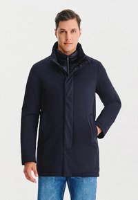 Granatowy ocieplany płaszcz z wysokim kołnierzem, ukrytym zamkiem błyskawicznym i zapięciami na zatrzask. Posiada boczne kieszenie oraz gładką, matową fakturę.