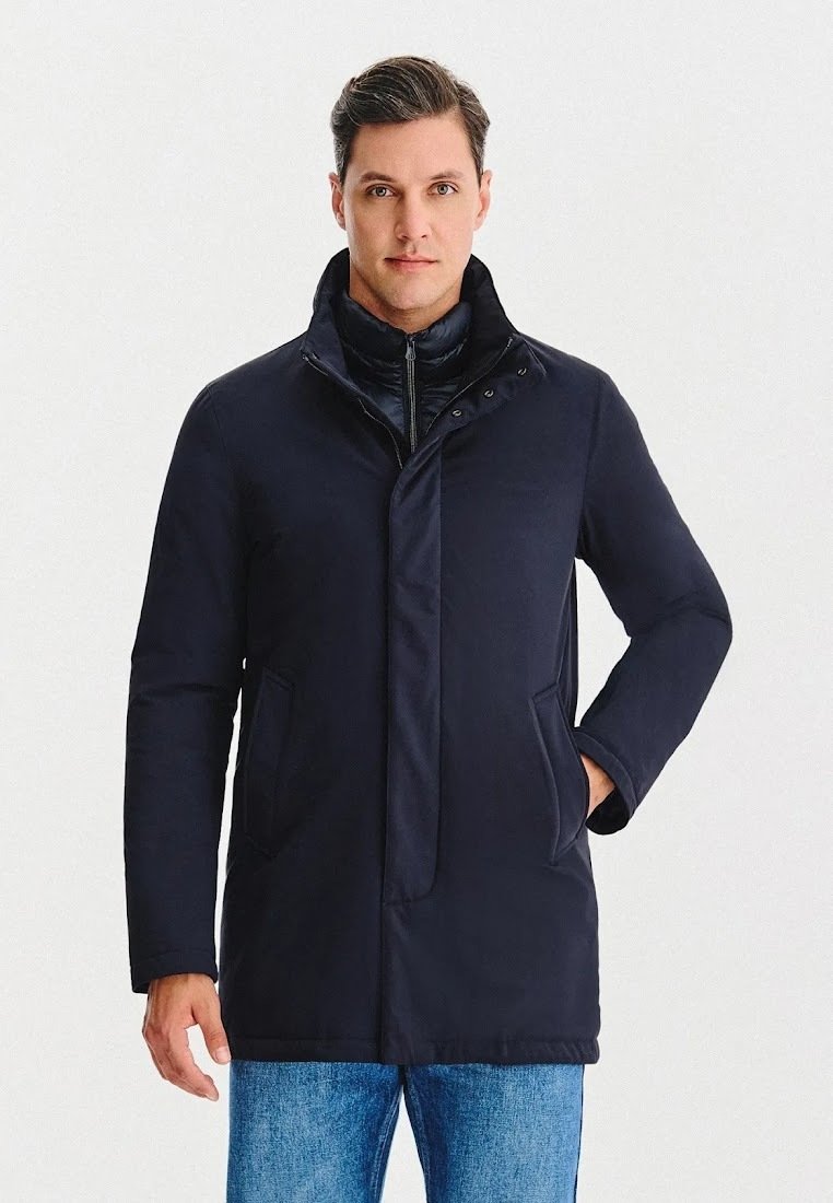 Granatowy ocieplany płaszcz z wysokim kołnierzem, ukrytym zamkiem błyskawicznym i zapięciami na zatrzask. Posiada boczne kieszenie oraz gładką, matową fakturę.