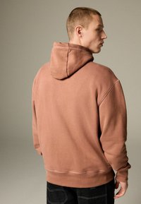 Brown hoodie with a drawstring hood, featuring a relaxed fit and visible stitching details. Worn with dark denim, plain background.